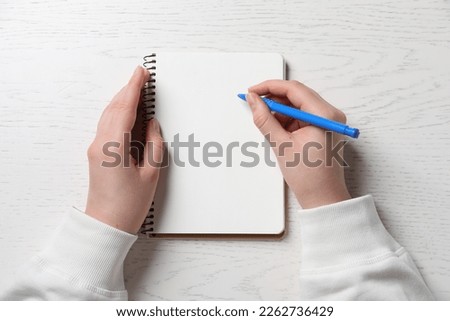 Woman writing in notebook with pen at white wooden table, top view
