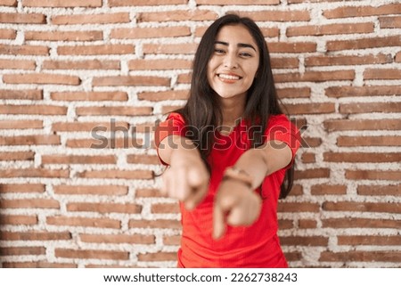 Young teenager girl standing over bricks wall pointing to you and the camera with fingers, smiling positive and cheerful 