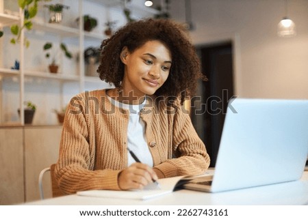 Confident African American young woman with stylish afro hairstyle, smart student studying, taking notes on notepad watching training courses on laptop. Online business or education concept Royalty-Free Stock Photo #2262743161
