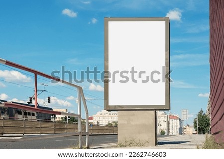 Empty vertical advertisement billboard mounted on urban sidewalk next to tram and bus stop against blue sky