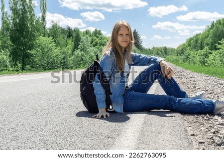 A young girl with a backpack is sitting on the side of the road waiting for vehicles. The concept of travel, hitchhiking, stupidity, vacation. High quality photo