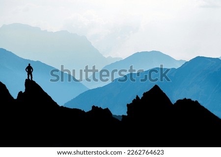 One Man reaching summit after climbing and hiking enjoying freedom and looking towards mountains silhouettes panorama during sunrise. Royalty-Free Stock Photo #2262764623