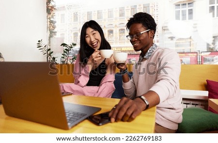 Happy multiethnic millennial friends sit in coffeeshop looking at laptop screen see pictures together.