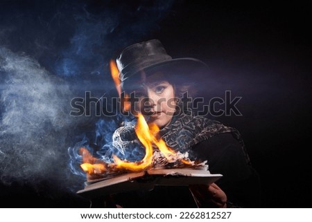 Portrain of witch girl with fier on magic Book. Woman in hat conjuring, making witchcraft over spooky dark black magic background. Model posing in Halloween party. Author writer burns manuscript