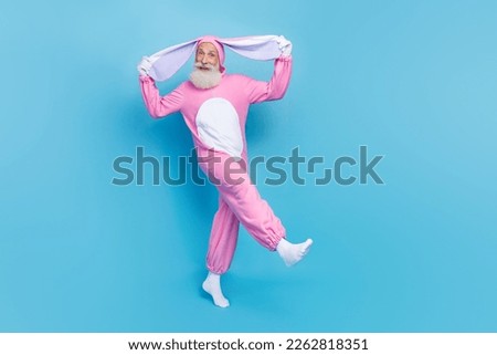 Full length photo of funky carefree retired guy wear pink hare sleepwear walking holding ears isolated blue color background