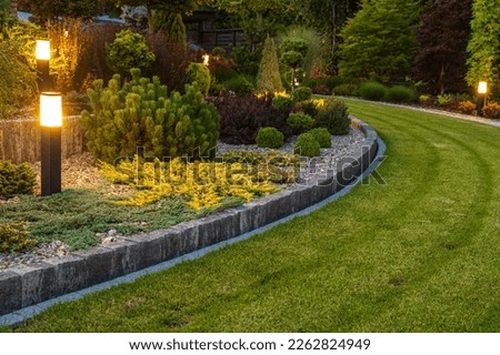 Professionally Landscaped Backyard Garden with Evenly Mowed Lawn and Trimmed Shrubs Illuminated with Outdoor Bollard Lamps. Royalty-Free Stock Photo #2262824949