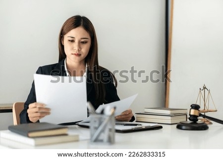 Business lawyer woman holding business contract to reading while sitting to working on the table with brass scales and justice hammer in law firm office.