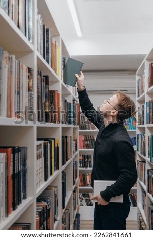 young adult man stands in library in middle of shelves and takes book. reading and recreation, education on university campus, free study of exact sciences, philosophy. bookstore. student 