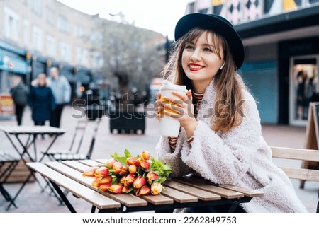 Stylish young smiling woman enjoying coffee from a reusable cup, listening to music, podcast using wireless earphones in a street cafe. Fresh tulips bouquet on the table. Springtime street fashion
