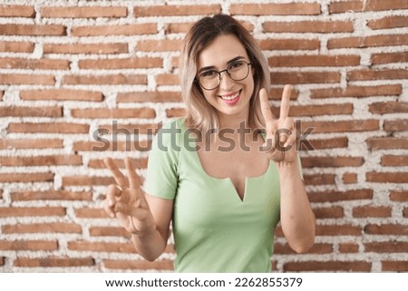 Young beautiful woman standing over bricks wall smiling looking to the camera showing fingers doing victory sign. number two. 