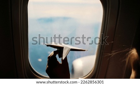 close-up. Silhouette of a child's hand with small paper plane against the background of airplane window. Child sitting by aircraft window and playing with little paper plane. during flight on airplane Royalty-Free Stock Photo #2262856301