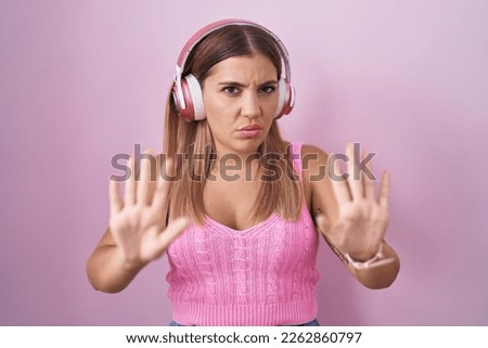 Young blonde woman listening to music using headphones moving away hands palms showing refusal and denial with afraid and disgusting expression. stop and forbidden. 
