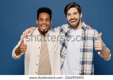 Young two friends happy fun cool men 20s wear white casual shirts together looking camera put hand on shoulder show thumb up isolated plain dark royal navy blue background. People lifestyle concept