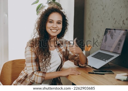 Young smiling happy cool fun smart successful employee business woman of African American ethnicity wear casual shirt look camera show thumb up sit work at office desk with laptop pc computer indoors