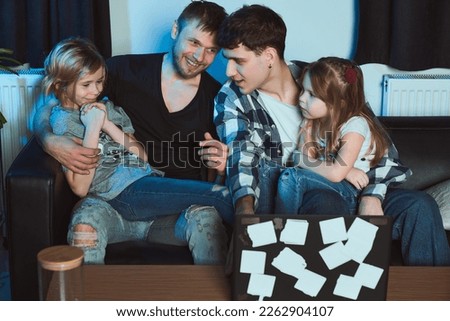 Staged photo. Homosexual couple and their children, two cute girls, at home.  An everyday evening after work. The whole family settled down on the couch. The girls sit on their parents' laps.         