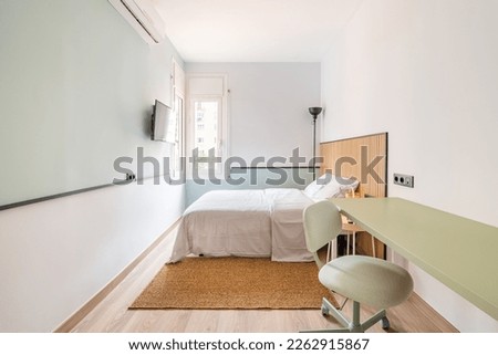 Narrow long room with bed, on opposite wall is TV for viewing watchable content. In corner is floor lamp with black shade for soft lighting at night. On floor is soft warm brown carpet. Royalty-Free Stock Photo #2262915867