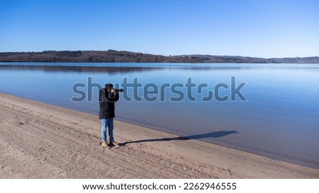 In the photograph a photographer is seen taking photos of a beach landscape with a blue sky and a calm lake.