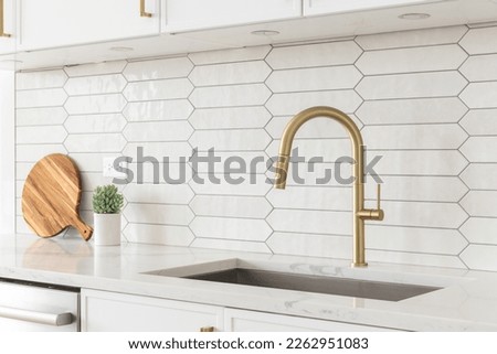 A beautiful kitchen faucet detail with white cabinets, a gold faucet, white marble countertops, and a brown picket ceramic tile backsplash. Royalty-Free Stock Photo #2262951083