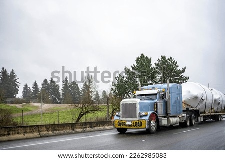 Blue classic big rig semi truck with oversize load sign on the bumper transporting oversized covered cargo on step down semi trailer running on the highway road with wet surface at raining weather