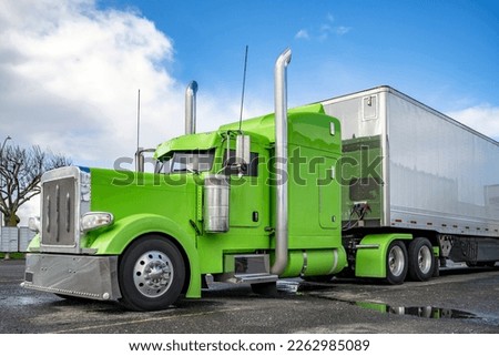 Power green classic American Idol big rig semi truck tractor with shiny chrome parts and high vertical exhaust pipes and dry van semi trailer standing on the parking lot waiting for the next load Royalty-Free Stock Photo #2262985089