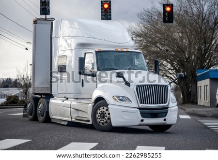 Commercial big rig classic white industrial grade long haul professional semi truck transporting cargo in dry van semi trailer turning on the city street crossroad with traffic light and crosswalk Royalty-Free Stock Photo #2262985255