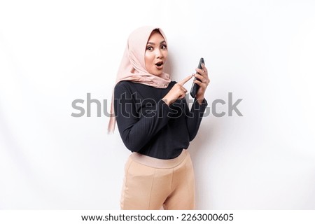 Shocked Asian woman wearing hijab, holding her phone, isolated by white background