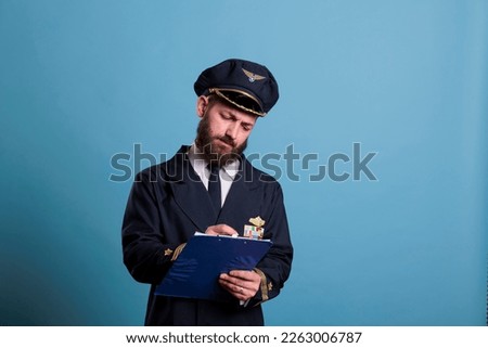 Airplane captain holding clipboard, concentrated pilot writing papers. Aircraft aviator in professional aviation uniform filling form in airport front view, studio medium shot on blue background