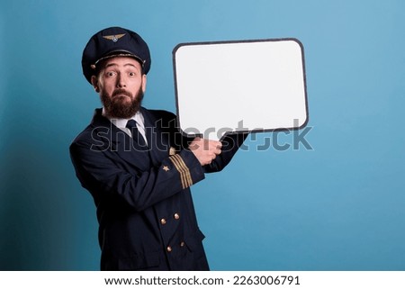 Surprised airplane pilot holding white empty speech bubble with copy space, aviator with communication frame mock up. Plane captain in uniform standing with blank message balloon