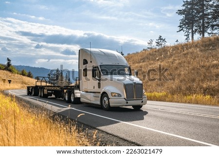 Industrial white long haul big rig high extended cab semi truck tractor transporting empty flat bed semi trailer running on the narrow winding highway road to warehouse for the next load Royalty-Free Stock Photo #2263021479