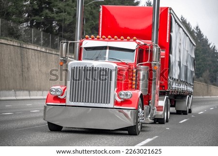 Classic big rig red semi truck tractor with chrome high exhaust pipes and truck driver sleeping compartment transporting cargo in covered semi trailer running on the road with wall on the side