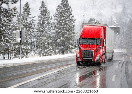 Red big rig commercial semi truck transporting cargo in dry van semi trailer running on the wet turning road with winter forest at snowing weather during a snow storm near Shasta Lake in California Royalty-Free Stock Photo #2263021663