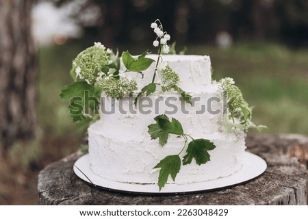 Three-tiered white wedding cake made of cream and biscuit stands on a stump in a summer forest and decorated with branches of greenery and flowers