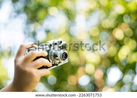 film camera in the hands of a girl taking a picture. Green trees bokeh background, woman taking photo with camera, selective focus, soft focus.