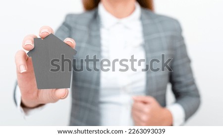 Businesswoman in gray suit holding colored paper house in one hand. Showing Important Informations. Executive Displaying Late News. Woman Showing Recent Updates.