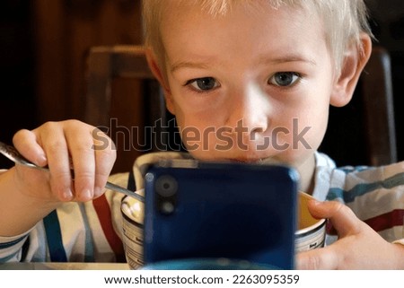 Little Boy using Phone while eating Soup during Lunch or Dinner at the Table. Child eating Food and watch Cartoon on Smartphone because Nomophobia or Attention Deficit Hyperactivity Disorder ADHD .