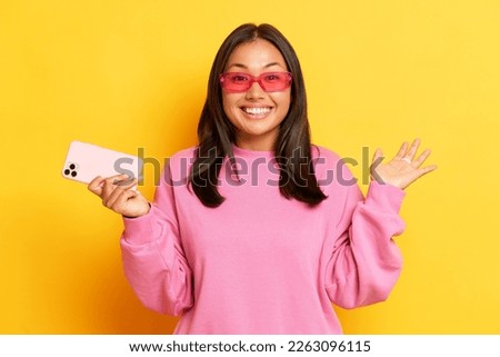 Positive charming brunette woman with joyful expression holds modern cellular smiles toothily wears stylish transparent eyewear and pink jumper poses over yellow background