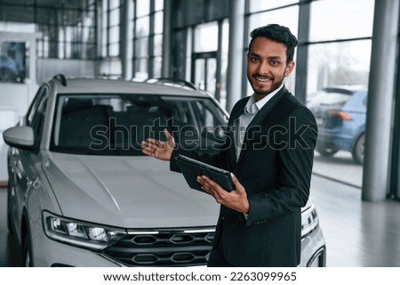 Holding graphic tablet. Handsome indian man in suit is in the car showroom with the vehicle indoors.