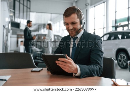 Man in headphones is sitting by table. Three people are working together in the car showroom.