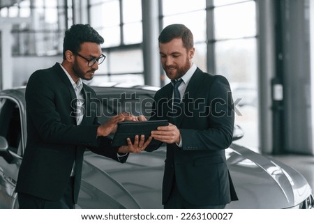 New plan. Standing near automobile with graphic tablet. Two businessmen are working together in the car showroom.