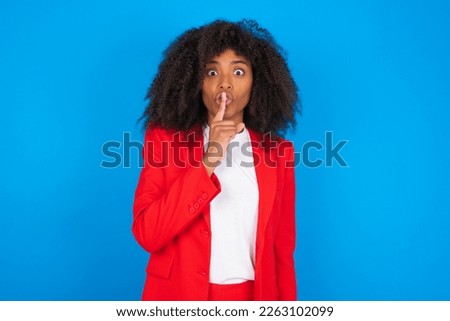 young businesswoman with afro hairstyle wearing red over blue wall makes hush gesture, asks be quiet. Don't tell my secret or not speak too loud, please!