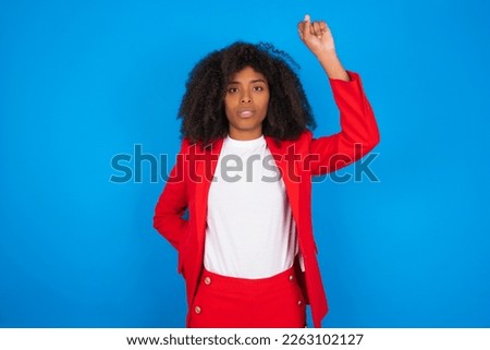 young businesswoman with afro hairstyle wearing red over blue wall feeling serious, strong and rebellious, raising fist up, protesting or fighting for revolution.