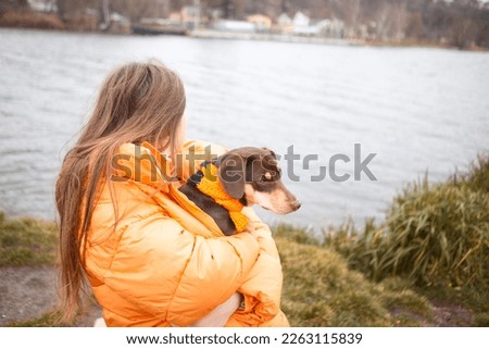 
A beautiful teenage girl in an orange jacket hugs and warms a dachshund dog on the river bank