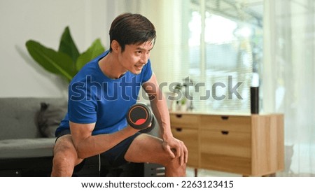 Sporty young man exercising with dumbbell in cozy home interior. Healthy lifestyle and fitness concept