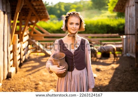 Beautiful young rural girl in medieval dress and bonnet hat posing on a ranch with jug of milk. Pretty blonde model in countryside art photo.  Royalty-Free Stock Photo #2263124167