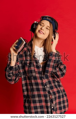 Young beautiful positive girl in checkered shirt and cap listening to music in headphones with player over red studio background. Concept of youth, beauty, lifestyle, emotions, facial expression. Ad