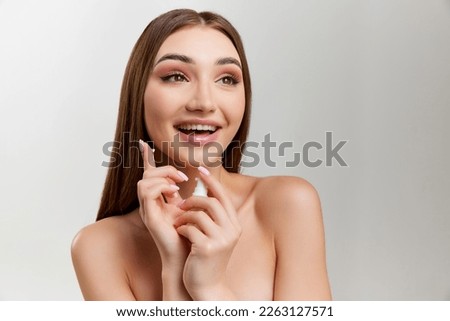 Smiling beautiful young girl with healthy, well-kept skin posing with face cream over grey studio background. Concept of natural beauty, skin care, cosmetology, cosmetics, health, plastic surgery