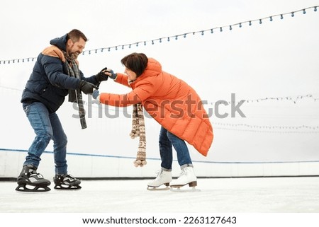 Middle-aged people, couple, having fun on outdoor ice-rink in winter day. Man teaching woman to skate. Concept of leisure activity, winter hobby and sport, vacation, fun, relationship, emotions.