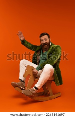 Excited bearded man in jacket waving hand while sitting on rocking horse on red background