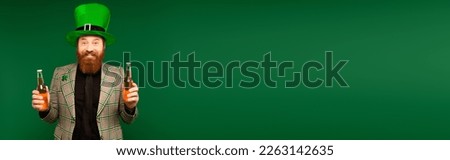 Positive bearded man in hat holding bottles of beer during saint patrick day isolated on green, banner