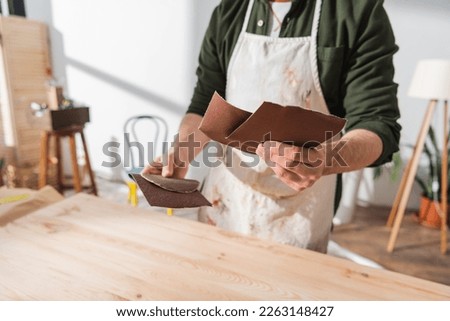 Cropped view of blurred craftsman in apron holding sandpaper while working with wooden board